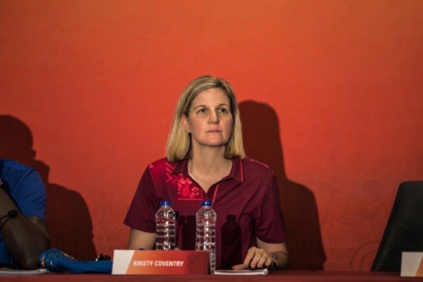 International Olympic Committee President Kirsty Coventry seated at a press conference table during Dakar 2026 wearing a maroon polo shirt with the Olympic rings in front of a red backdrop with sponsor logos  trybalnews