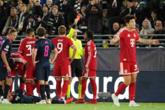 Michael Olise of FC Bayern Munich, wearing the red Bayern kit with dreadlocks, receives a red card from the referee during the UEFA Champions League 2026/27 League Phase MD2 match between Viking FK and FC Bayern Munich at Viking Stadion on October 13, 2026, in Stavanger, Norway. Teammates including Harry Kane (number 9) look on as a Viking FK player lies injured on the pitch.