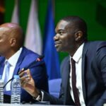 Samson Adamu in a dark suit gesturing while speaking into a microphone at a conference table with CAF President Patrice Motsepe in a blue suit sitting next to him  trybalnews