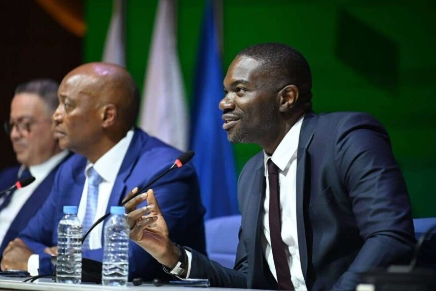 Samson Adamu in a dark suit gesturing while speaking into a microphone at a conference table with CAF President Patrice Motsepe in a blue suit sitting next to him  trybalnews