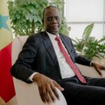 Prime Minister of Senegal Ahmadou Alhaminou Mohamed Lo in dark suit and red tie sitting in beige armchair next to Senegal national flag in bright office with green plants  trybalnews