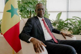 Prime Minister of Senegal Ahmadou Alhaminou Mohamed Lo in dark suit and red tie sitting in beige armchair next to Senegal national flag in bright office with green plants  trybalnews