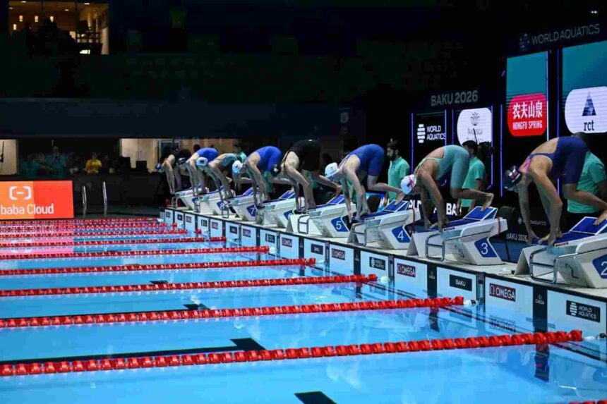 Eight female swimmers in colored swimsuits and swim caps bent over starting blocks ready to dive into Olympic pool with red lane dividers at World Aquatics Baku 2026 competition with Omega and World Aquatics branding visible  trybalnews