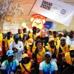 IOC President Kirsty Coventry smiling in group photo with Dakar 2026 volunteers wearing yellow shirts and orange caps with Dakar 2026 flag and lion mascot in background at airport  trybalnews