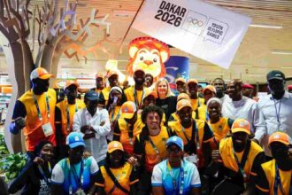 IOC President Kirsty Coventry smiling in group photo with Dakar 2026 volunteers wearing yellow shirts and orange caps with Dakar 2026 flag and lion mascot in background at airport  trybalnews